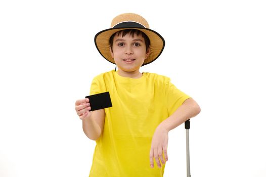 Isolated portrait on white background of a Latin American happy teenage traveler boy in yellow t-shirt and straw hat, with suitcase, showing a blank black plastic card with free ad space on camera