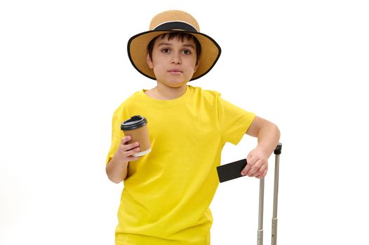 Isolated portrait on white background of teenage traveler boy, wearing straw hat and yellow t-shirt, looking at camera, posing with black plastic credit card, suitcase and takeaway drink in paper cup