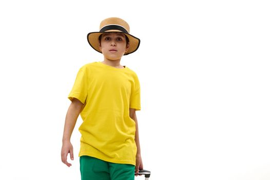 Adorable Caucasian teenage boy in straw hat and bright summer wear, walking with suitcase over white background with copy ad space. Cheerful kid going for summer vacations. Trip Travel Journey concept