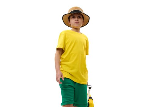 Charming teenage boy in yellow t-shirt, green summer shorts and straw hat, with suitcase, going for weekend getaway, white background. Travel destinations, journey, air trip concepts. Copy ad space