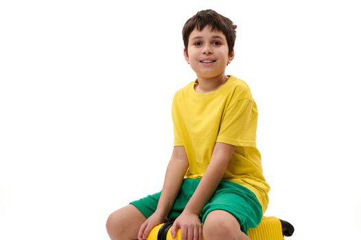 Happy teenage boy in yellow t-shirt and green shorts, sitting on a suitcase and ready for travelling. Child tourist packs clothes into a suitcase for travel. Travel and adventure concept