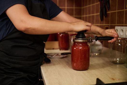 Close-up. Selective focus on a chef's hands using seamer or special seaming key, close lids of jars with freshly canned tomato sauce, juice or passata. Canning homegrown organic tomatoes for winter