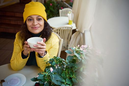 Close-up of stunning Middle Eastern woman in yellow jacket and hat, taking sip of hot drink, warming her hands with a cup of tea or coffee, smiling at camera, enjoying outdoor coffee break on fall day