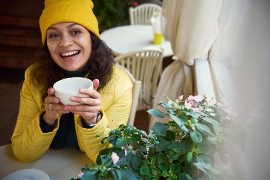 Beautiful dark-haired Latin American woman in yellow hat and jacket, sits at table with cup of hot drink in hands, smilesa toothy smile looking at camera, chilling in a coffee shop with rustic design