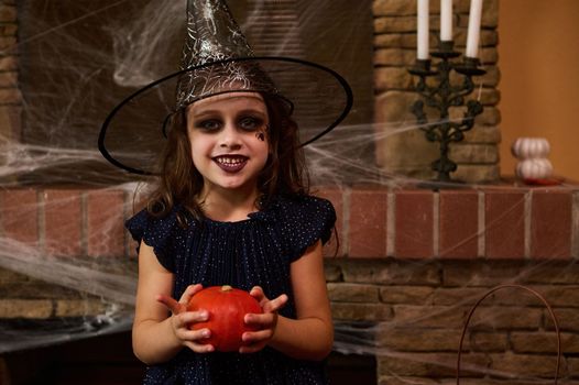 Little girl in witch dress and wizard hat with art make-up on her face, holding a pumpkin, smiling a toothy smile looking at the camera, sitting against a fireplace, decorated with spider web, candles
