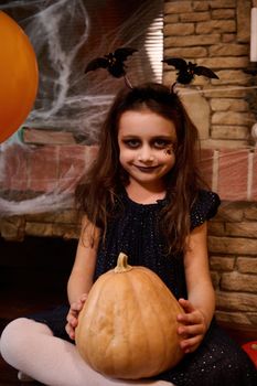 Little girl in black dress and a smokey eyes makeup, smiles looking at camera, making Halloween decoration at home, drawing scary face on pumpkin and enjoying holiday preparation.