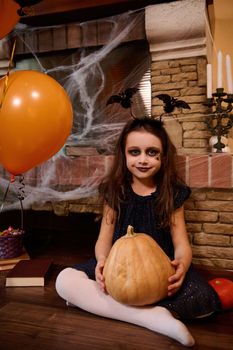 Adorable stylish little girl with smokey eyes makeup, wearing black dress and hoop with bats, holding a Halloween pumpkin, sitting by a spider web covered fireplace and air balloons, smiling at camera