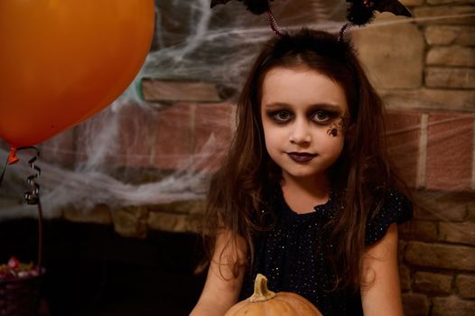 Charming Caucasian little girl, looking like a sorceress in witch carnival costume and smokey eyes gothic makeup, smiles looking at camera, enjoying Halloween preparations. Adorable enchantress