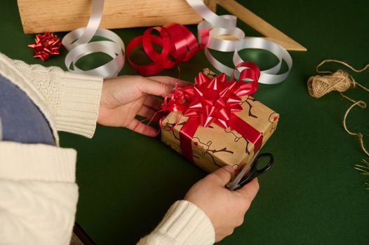 Overhead view of woman's hands tying bow from shiny red ribbon while decorating a Christmas gift. Wrapping. Packaging presents for New Year or any other celebration event. December 25th. Boxing Day.