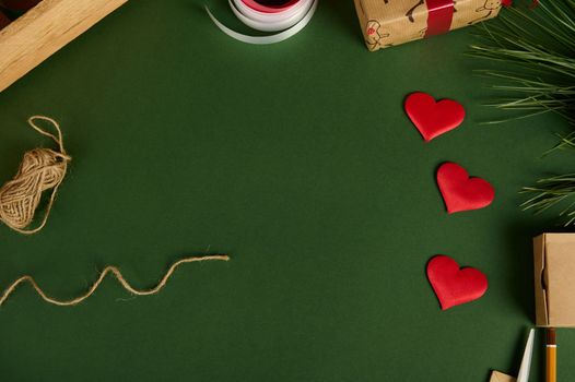 Top view of red decorative hearts, linen rope and wrapping materials on a green background with copy advertising space. Boxing day. Christmas mockup. Handwork art. Diy presents. Flat lay. Still life