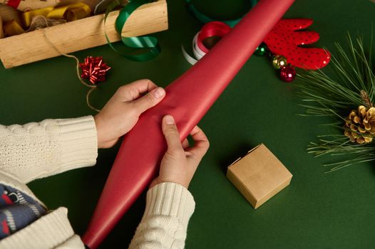 Top view woman's hand rolling out a red gift paper while, choosing wrapping decorative materials, over green background. Golden envelope, tied bow and tapes on wooden crate. Packing Xmas diy presents