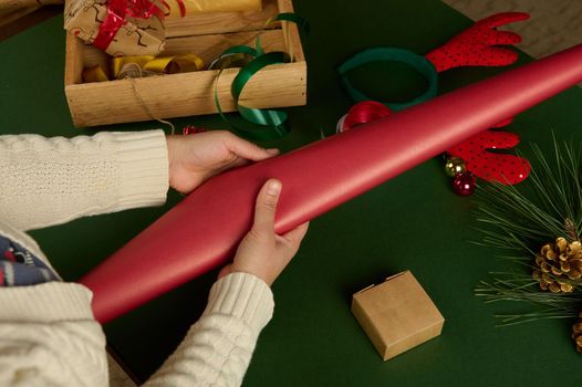 Overhead view woman's hands hold gift wrapping paper of red color while packing Christmas New Year's presents, on green background. Festive decorations in wooden crate.Boxing Day. Xmas. December 25th