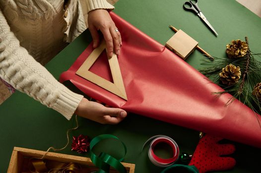 Close-up. View from above of woman's hands using wooden triangular ruler, calculate the amount of wrapping paper for packing Christmas or New Year's gifts, at green background. Boxing Day. Copy space