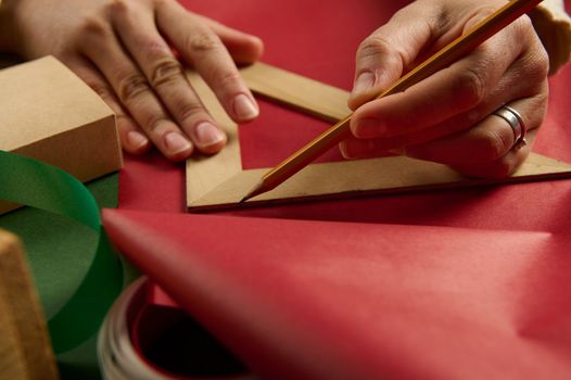 Close-up woman's hands using a wooden triangular ruler to draw on red gift paper, calculating the amount of packaging materials needed to wrap gifts for Christmas, New Year or any other holiday event
