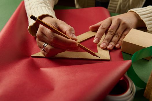 Details: woman's hands with a wooden crayon and triangular ruler drawing on a red wrapping paper, calculating the amount of it for wrapping presents for Saint Valentine's Day, Christmas or New Year's