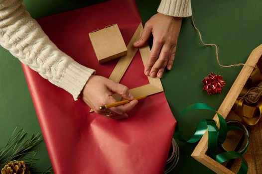 Top view female hands using a wooden pencil and triangular ruler drawing on a red wrapping paper, calculating the amount of it for wrapping presents for Saint Valentine's Day, Christmas or New Year's