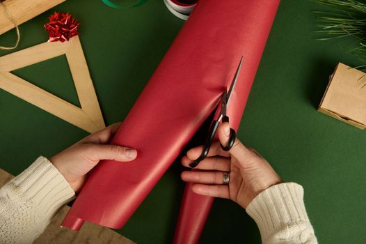 Top view of a woman cutting the red decorative wrapping paper with scissors, for packing gifts, over a green background. Boxing Day. Christmas and New Year's preparations. Diy presents. Handwork art