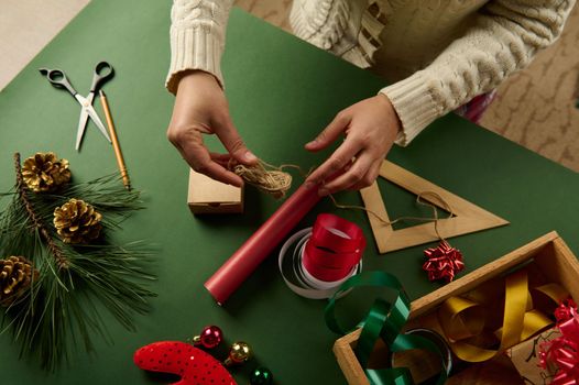 Top view of a woman's hands holding a linen rope for tying presents over a red gift wrapping paper. Packaging gifts for Christmas or any other celebration events. Boxing Day. New Year's preparations