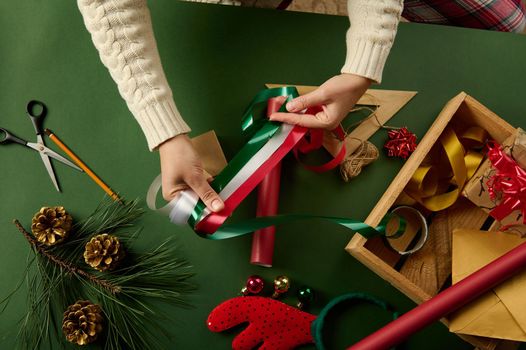 Top view woman's hands holding red, white and green shiny decorative ribbon over a background with wrapping materials, for packing gifts for Christmas, New Year or other celebration event. Boxing Day