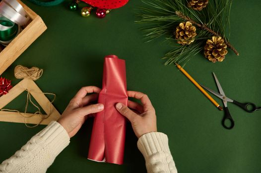 Close-up. Top view of a woman packaging a small present in red decorative paper over a green background with wrapping materials and golden pine cones as Christmas decor. Diy presents. Boxing Day