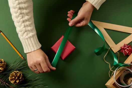 Woman's hands wrapping Christmas gift box, close up. Unprepared presents on green background with decor elements and items. Christmas or New year diy packing Concept. Boxing Day. Copy ad space