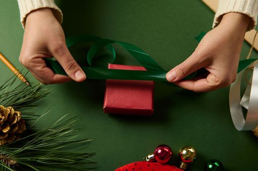 Top view of the hands of a woman in beige warm sweater, holding a shiny decorative ribbon while wrapping Christmas diy presents at green background with copy space. Boxing Day. December 25th