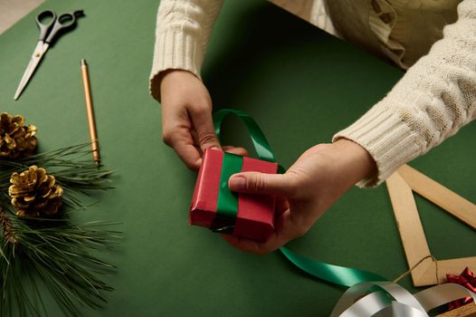 Close-up woman tying up present, wrapped in red paper with green decorative ribbon. Packing Christmas and New Year gifts. Happy holiday present, surprise. Magic time. Celebration winter event