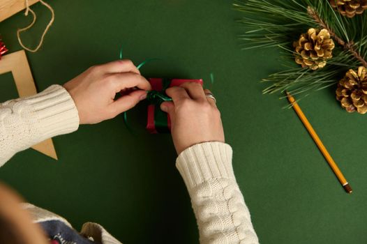 Close up. View from above of woman in warm beige sweater, decorating Christmas presents, wrapped in red gift box, making bow from green shiny ribbon. Boxing Day. Xmas and New Year celebration concept
