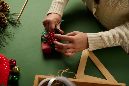 Close-up woman wrapping a small Christmas present in red gift paper and tying up with green decorative tape, tying a bow. Boxing Day. Packing presents. Celebrating winter events. New Year preparations