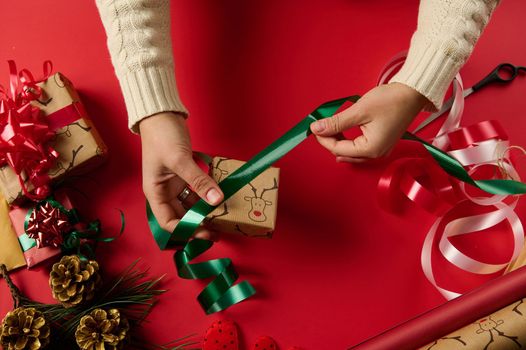Christmas wrapping idea, xmas wrapping workspace. Female hands wrap gift box in paper and tying bow on red surface. Close Up woman wraps a present, decorates with shiny green ribbon and red tied bow