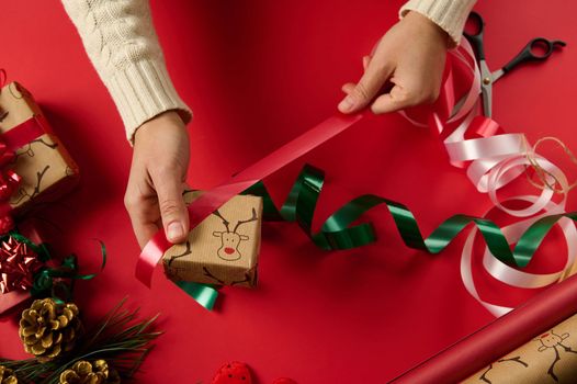 Christmas wrapping idea. Female hands wrap gift box in gift paper with deer pattern, tying bow on red surface. Close Up woman wraps a present, decorates with shiny red ribbon. Xmas preparations