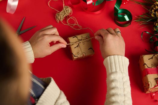 Close up. Overhead view woman in warm beige sweater, decorating Christmas presents, wrapped in deer pattern gift paper, making bow from linen rope . Boxing Day. Xmas and New Year celebration concept