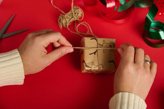 Top view of woman tying up a Christmas gift with a linen rope, over a red background, surrounded by festive ornaments, wrapping papers, ribbons. Packing presents. Boxing Day. New Year preparations