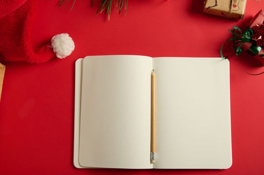 Still life with a wooden pencil on an open notepad or agenda with blank paper sheets and copy advertising space for promotional text, isolated on a red background with Christmas items and Santa's hat