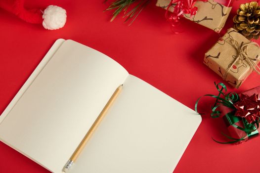 Top view of a wooden crayon on an open notepad or New Year agenda with blank paper sheets, copy advertising space for promotional text, isolated on red background with Christmas items and Santa's hat