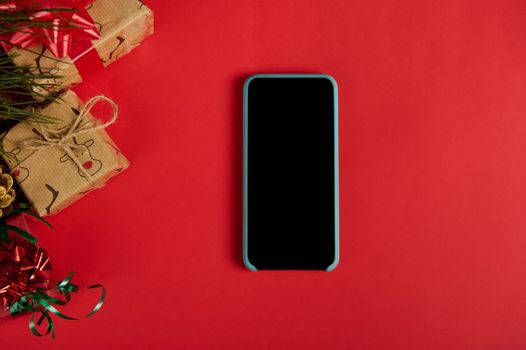 Flat lay. Still life with a modern smartphone with blank screen, next to Christmas and New Year's gift boxes and golden pine cones as a festive ornament, isolated over a red background. Copy ad space
