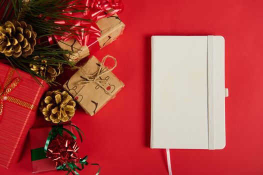 Flat lay. A white notepad or agenda book next to Christmas gift boxes, decorated with ribbons and bows and golden pine cones with fir-tree branch, isolated over red background. Copy ad space. Mockup
