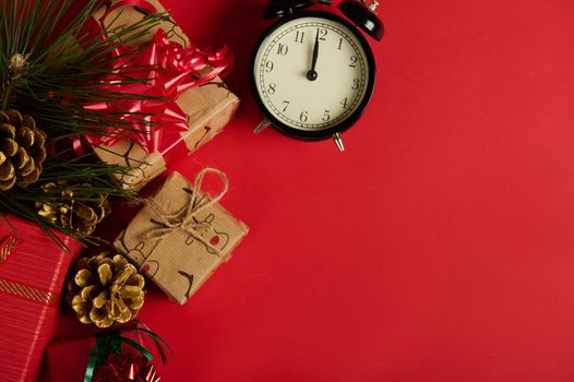 Still life. Alarm clock with midnight on dial next to gifts and fir branch with golden pine cones - as a Christmas ornament, isolated on red background with copy ad space. Mockup. New Year's event