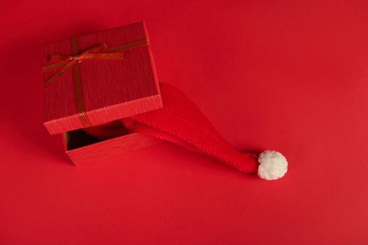 Top view of half open stylish red gift box, tied up with a red bow, with a Santa hat inside it, isolated over bright red background with copy ad space. New Year and Christmas preparations. Boxing Day