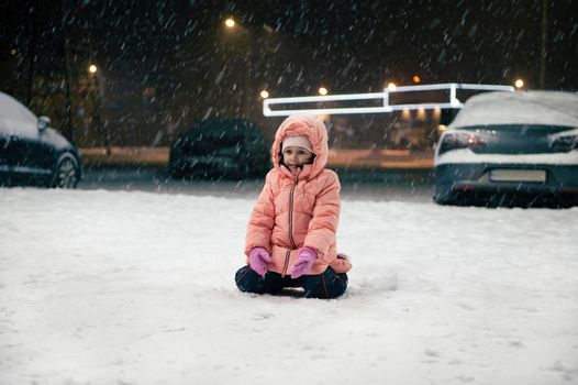 Happy Caucasian child, preschooler little girl in warm winter pink overalls sits on a snowy street, enjoying the first snow during an evening walk. Winter. Snow covered city. Childhood.