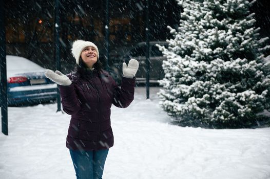 Charming cheerful multi-ethnic woman wearing wool mittens and warm clothes, looking up the evening sky, enjoys the snowfall in the city