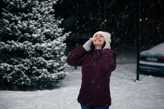 Happy cheerful multi-ethnic woman in warm clothes, having fun outdoor, smiling a toothy smile looking up the sky, enjoys a snowfall on a snow covered city. Winter holidays. Merry Christmas concept