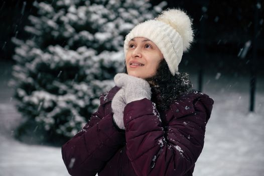 Close-up portrait of charming dark-haired multi-ethnic woman in warm clothes, white wool hat and mittens in the snow covered nature. Snowfall. Winter holidays. Happy New Year. Mmerry Christmas concept