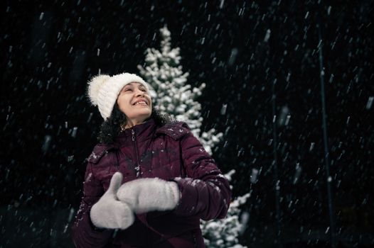 Portrait of a delightful beautiful multi ethnic woman in warm clothes looking up to the sky, enjoying christmas atmosphere outdoors on snowy evening. Snowfall. Winter holidays. Merry christmas concept