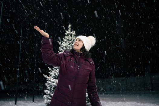 Delightful multi-ethnic woman in warm clothes, catching snowflakes on her hand while walking down the city street, on a snowy evening. Winter. Snowfall. Merry Christmas and Happy New Year concept