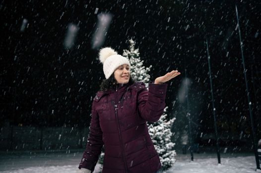 Charming happy woman in warm clothes, catching snowflakes on her hand while walking down the city street, on a snowy evening. Winter. Snowfall. Merry Christmas and Happy New Year concept