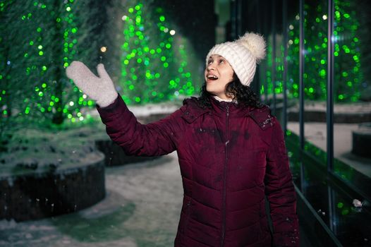 Pretty multi-ethnic woman in warm clothes, catches snowflakes, enjoys walking down the city street, lightened by garlands on a snowy winter night. Winter holidays. Snowfall. Merry Christmas