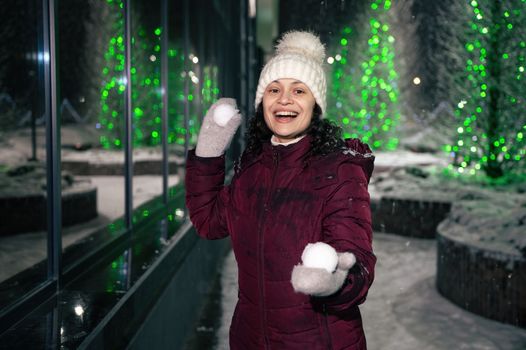 Beautiful multi-ethnic woman wearing warm clothes, laughing, smiling, having fun in a city street, illuminated by garlands, playing with snowballs on a winter snowy evening. Christmas holidays concept
