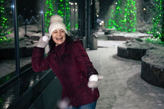 Attractive Hispanic woman wearing warm clothes, laughing, smiling, having fun in a city street, illuminated by garlands, playing with snowballs on a winter snowy evening. Christmas holidays concept