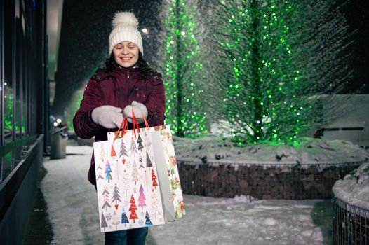 Multi-ethnic pretty woman smiling a beautiful toothy smile, walking with shopping bags on the street, illuminated by garlands at snowy winter night. Merry Christmas and Happy winter holidays concept